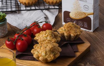 Fotografia em tons escuros de uma tábua de madeira quadrada com um pano marrom, tomates cereja e três tortinhas de legumes. Ao lado tem uma embalagem de farelo de aveia, e um pano branco com uma grade preta por cima, mais tortinhas apoiadas.