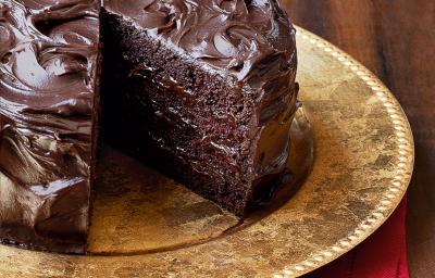 Fotografia de um bolo marrom de chocolate com uma fatia faltando, em um prato dourado em cima de um guardanapo de tecido vermelho em uma mesa de madeira escura.