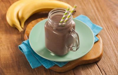 Fotografia em tons escuros, em uma bancada de madeira de cor marrom. Ao centro, uma tábua de madeira marrom contendo um pano azul. Em cima, há um prato verde e redondo contendo um copo com o milkshake. Ao fundo, há 2 bananas.