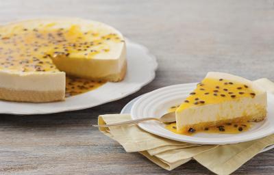 Fotografia em tons de amarelo e branco, com prato branco contendo uma torta de maracujá e ao lado um prato de sobremesa com uma porção da torta e uma colher de sobremesa, sobre guardanapo amarelo e outro prato, tudo sobre bancada cinza.