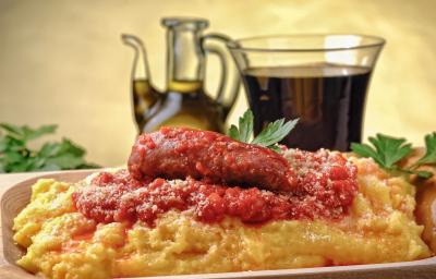 Fotografia de uma polenta com molho de tomate, cebolas e linguiça, que está dentro de um prato fundo de madeira. Por cima, queijo ralado e algumas folhas de salsinha. No fundo, muito azeite em um recipiente de vidro na frente de uma parede amarela.