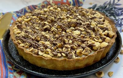 Foto da receita de Torta Nocciolíssima. Observa-se uma torta inteira decorada com avelãs e creme de chocolate sobre um prato de cerâmica redondo preto.