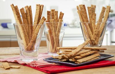 Fotografia em tons de pardo e vermelho de uma bancada de madeira marrom vista de frente, contém 3 copos transparentes e cada um contém biscoitos de cebolas em tiras. Ao lado uma travessa preta com mais biscoitos em tiras e abaixo um pano vermelho.