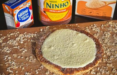 Fotografia em tons claros de uma tábua de madeira salpicada com farelo de aveia e tem uma panqueca de aveia com creme de ninho por cima e ninho salpicado. Atrás da tábua tem aveia e produtos zero lactose nestlé. Fotografia em tons claros de uma tábua de madeira salpicada com farelo de aveia e tem uma panqueca de aveia com creme de ninho por cima e ninho salpicado. Atrás da tábua tem aveia e produtos zero lactose nestlé.