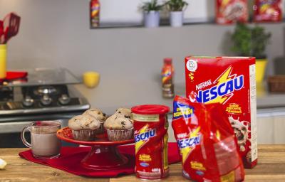 Foto vista de frente para um bancada com produtos Nescau à direita, mais ao centro um prato vermelho alta com quatro Muffins Crocantes de Nescau e à esquerda uma caneca com leite e Nescau.