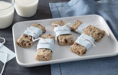 Foto em tons de azul claro da receita de barrinha de iogurte nesfit e aveia servida em 5 porções sobre uma régua de porcelana branca sobre uma mesa cinza com um pano azul ao fundo além de dois copos com iogurte