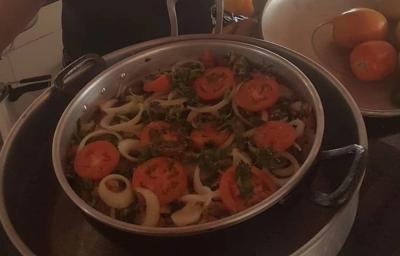 Fotografia de uma panela grande com moqueca com tomates, pimentões e cebolas em rodelas por cima. A panela está dentro de uma outra panela maior.