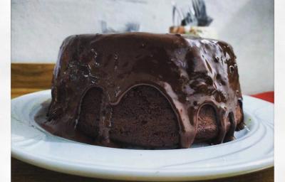 Fotografia em tons de preto em uma mesa de madeira, um pano vermelho, um prato branco raso redondo com o bolo de chocolate com cobertura de chocolate em cima.