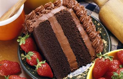 Fotografia de uma fatia de bolo com massa e recheio de chocolate, morangos por toda a parte em um prato desenhado de cor escura, apoiado em uma toalha de mesa azul e branca, quadriculada. Ao lado do bolo, dois potes e um rolo de massa.