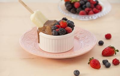 Fotografia em tons de rosa e branco de uma bancada rosa vista de frente, ao centro um prato redondo rosa e por cima um potinho branco com frutas vermelhas e um picolé mergulhando na calda de chocolate. Ao fundo um prato transparente com frutas vermelhas.