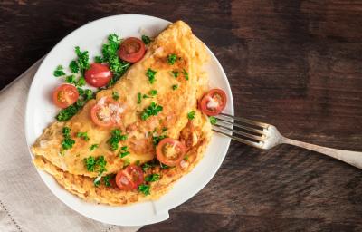 fotografia em tons de marrom, branco, amarelo e vermelho de uma bancada marrom vista de cima. Contém um prato redondo branco com uma omelete, e por cima pedaços de tomates e salsas