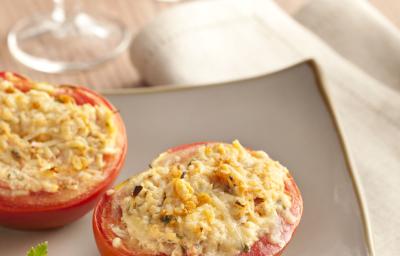 Fotografia em tons de vermelho e bege, em uma mesa de madeira a receita de tomate recheado é servida em um prato branco, e para decorar  um guardanapo branco com taças ao fundo