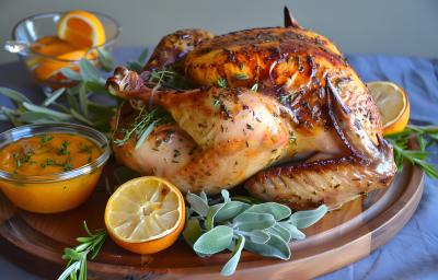 Fotografia de um frango inteiro assado com mel e laranja sobre uma tábua de madeira redonda. Por cima da tábua tem alguns ramos de tomilho, laranjas na metade e um molho laranja dentro de um recipiente de vidro pequeno.