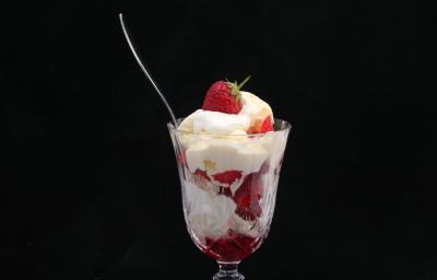 Fotografia de uma taça de vidro com merengue e morangos, de forma intercalada. E dentro do recipiente, uma colher de sobremesa apoiada, sobre um fundo preto.