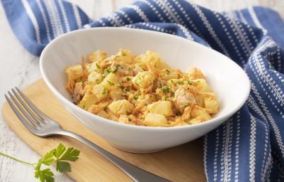 Fotografia em tons de azul em uma bancada de madeira com um pano listrado azul ao lado. No centro, um prato fundo branco com a salada de batata, atum, cebola e salsinha.