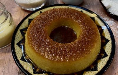 Foto em tons de bege da receita de pudim de tapioca servida em um prato de porcelana amarelo com adornos em marrom. Ao fundo, uma xícara de tapioca granulada, um coco cortado ao meio e um vidro com leite MOÇA