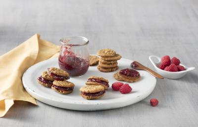 fotografia em tons de cinza, bege e vermelho de uma bancada cinza vista de cima. Ao centro um prato branco redondo com um pote transparente com geleia e em volta vários Whoopies recheado com a geleia. Do lado esquerdo um pano bege.