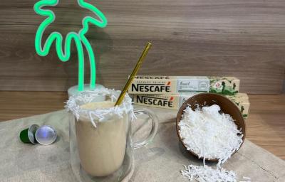 Fotografia em tons de verde com uma caneca de vidro ao centro. Dentro da caneca existe uma mistura de café gelado com leite de coco. Ao lado existe um pote marrom cheio de coco ralado e ao fundo duas caixas de cápsulas NESCAFÉ