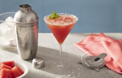 Foto em tons de vermelho da receita de margarita de melancia servida em uma taça de vidro sobre uma mesa de mármore onde estão uma coqueteleira cromada, além de um pano vermelho, uma mola para mistura, um copo com gelo e pedaços de melancia em cubos.