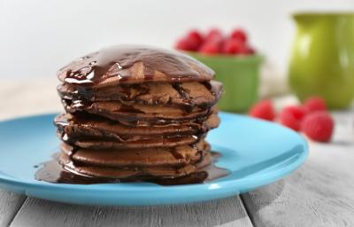 Fotografia de algumas panquecas de chocolate empilhadas em um prato azul claro, e sobre as panquecas tem uma calda de chocolate escorrendo. Ao fundo em algumas framboesas dentro de um recipiente verde, e espalhadas sobre a mesa de madeira branca.