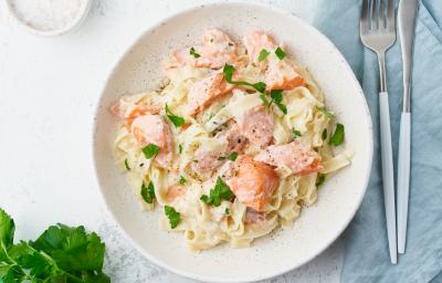 Fotografia em tons de branco, rosa, amarelo e verde de uma bancada branca vista de cima. Ao centro contém um prato redondo com macarrão, pedaços de salmão e salsa.