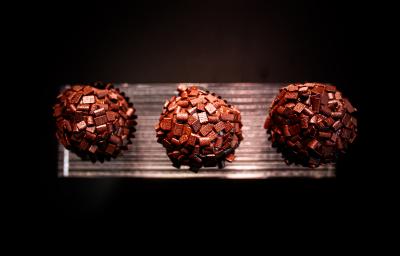 Fotografia vista de cima de três brigadeiros de tucupi reduzido recheados com cupuaçu e enrolados no chocolate amargo. Os brigadeiros estão em um apoio na cor prata, com o fundo escuro.