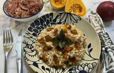 Foto da receita de risoto de carne seca e abóbora em tons de laranja servida em um prato de porcelana azul e branco. Ao lado, há talheres prateados e um guardanapo adornado, além de carne seca, abóbora e cebola ao fundo para decorar.