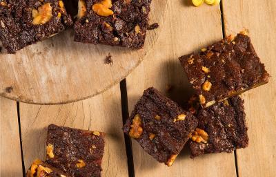 Fotografia em tons de amarelo em uma bancada de madeira com uma tábua de madeira clara com alguns brownies de chocolate em cima. Na bancada, alguns brownies espalhados e flores amarelas espalhadas na mesa para decorar.