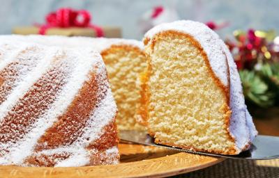 Fotografia de um bolo de massa branca de leite com um pedaço em destaque e cobertura de açúcar de confeiteiro, sobre um apoio reto de tom claro.