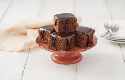 Fotografia em tons de chocolate e branco, ao centro boleira vermelha contendo pedaços de bolo de chocolate com calda de chocolate, ao lado pratos empilhados com 3 garfos por cima, guardanapo rosa, tudo sobre bancada branca.