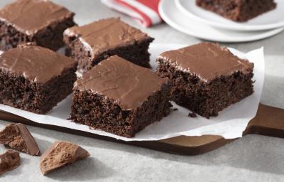 Fotografia em tons de marrom e branco de uma bancada branca  com uma tábua de madeira, sobre ela um papel manteiga com pedaços de bolo de chocolate zero. Ao fundo dois pratos brancos com uma fatia de bolo de chocolate e um paninho vermelho e branco.