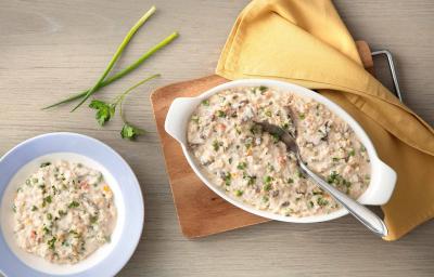 fotografia em tons de cinza e bege de uma bancada cinza vista de cima. Contém uma tábua de madeira com um recipiente oval branco com alças que contém o arroz  com creme, pedaços de carne e ervilhas por cima e ao lado um pano amarelo.