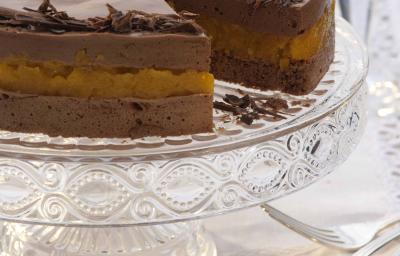 fotografia em tons de branco e marrom de uma bancada com panos branco, contém um suporte para servir sobremesas transparente com uma torta de mousse de chocolate com uma fatia retirada