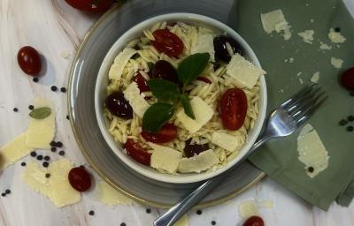 Foto da receita da salada de risoni servida em um bowl branco em cima de um prato de vidro transparente com um garfo prateado ao lado e um pano verde decorativo. O prato em cima de uma mesa clara de madeira com os ingredientes da salada como decoração.