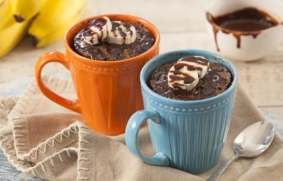 Fotografia em tons de beje, azul e laranja de uma bancada branca com um paninho beje, sobre ele um caneca azul e uma caneca laranja com bolo de caneca. Ao fundo um cacho de banana e um recipiente redondo com calda.