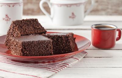 Fotografia em tons de vermelho em uma bancada de madeira clara com um pano listrado em branco e vermelho e um prato vermelho com três pedaços de bolo de cacau com coco. Ao lado, uma xícara vermelha de café e ao fundo, dois bules brancos.