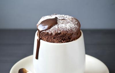 Fotografia de um suflê de café com NESCAFÉ e açúcar de confeiteiro polvilhado por cima. A sobremesa está em um recipiente de vidro branco alto em cima de um prato pequeno branco de sobremesa, e uma colher de café apoiada.