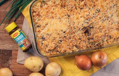 Fotografia em tons de amarelo e castanho claro com um refratário situado ao centro. Nele existe um gratinado de queijo na cor dourada, ao lado existe duas batatas e duas cebolas em cima de uma toalha de cor amarela.