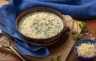 Fotografia em tons e marrom, bege e azul, com bowl marrom e bege ao centro contendo sopa com queijo e espinafre sobre prato marrom e tábua bege, ao entorno guardanapo azul, potinho com queijo e colher com aveia, tudo sobre bancada marrom.