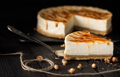 Fotografia de uma fatia de cheesecake com caramelo salgado por cima de uma espátula de prata, sobre uma mesa preta. No fundo tem o resto da cheesecake sobre um apoio preto.