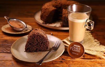 Fotografia em tons de marrom em uma bancada de madeira clara, um guardanapo de pano bege de palha, uma xícara em cima com o Café ao Leite Dolce Gusto. Ao centro, um prato redondo com um pedaço do bolo de chocolate em cima dele. Ao fundo, o bolo inteiro.