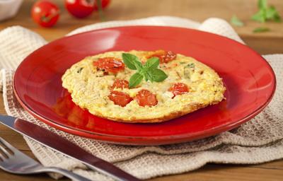 Fotografia em tons de vermelho em uma bancada de madeira, um pano bege, um prato vermelho redondo com a omelete com tomates-cereja em cima dele. Ao fundo, tomatinhos para decorar e ao lado, garfo e faca.