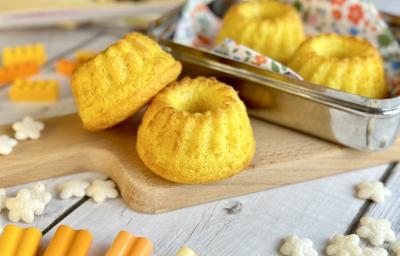 Fotografia em tons de laranja em uma bancada de madeira clara, uma lancheira ao meio com alguns bolinhos de milho dentro e em cima de uma tábua de madeira. Ao lado, brinquedos infantis.