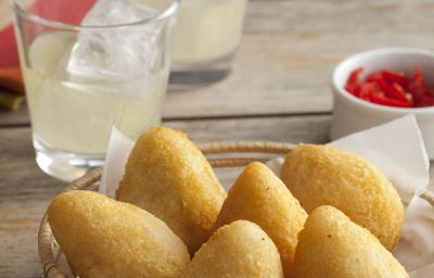 Fotografia em tons de dourado e grafite, com cestinha de vime ao centro contendo 6 coxinhas fritas, no lado direito potinho com pimentas vermelhas, ao alto dois copos com suco e gelo e um guardanapo vermelho e branco, tudo sobre bancada rústica.