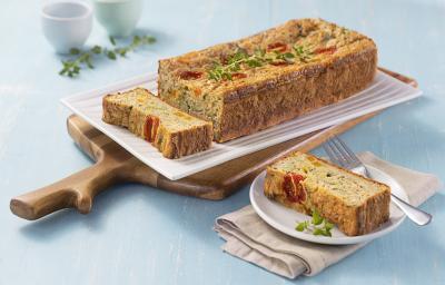 Foto da receita de Tortinha de Legumes. Observa-se uma superfície azul clara, com uma tábua de madeira e, em cima, um prato branco retangular com a torta de legumes inteira, também retangular. Ao lado, um prato branco redondo com um pedaço da torta.