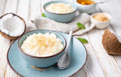 Foto da receita de Doce de Coco. Observa-se o doce em uma vasilha de cerâmica azul bebê, sobre um pires da mesma cor. Cocos cortados ao meio decoram a foto.