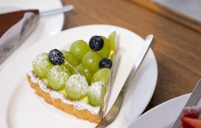 Fotografia de uma fatia de torta recheada com leite MOÇA e uvas verdes por cima, com três uvas roxas. Por cima da torta tem açúcar de confeiteiro polvilhado, e ao lado, um garfo sobre um prato branco, que está sobre uma mesa de madeira.