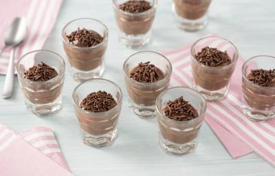 Fotografia em tons de azul e rosa claros de uma bancada azul clara com paninhos rosas com listras brancas, sobre eles nove copinhos de vidro redondos com brigadeiro de colher e granulados de chocolate.