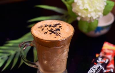 Fotografia de uma xícara de vidro com uma bebida com chocolate, Nescafé e leite. Ao lado, uma embalagem de Nescafé, e ao fundo, decoração de plantas e flores, sobre uma mesa preta.