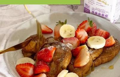 Foto em tons claros da foto da receita de french toast servida com bananas, morangos e uma calda sobre um prato de porcelana branco. Ao fundo, há uma embalagem de bebida vegetal de aveia natures heart